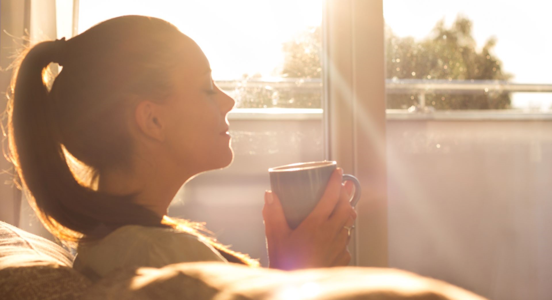Woman sitting on couch with coffee mug with early morning sunlight in window