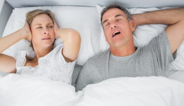 Woman laying in bed covering her ears next to snoring man