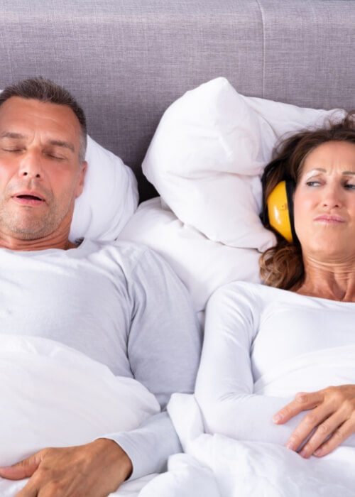 Woman in bed wearing headphones next to snoring man